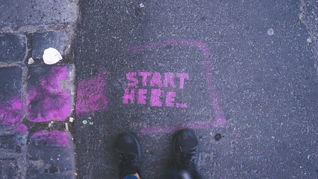 The words "start here" are painted on the pavement, with someone's feet standing before the words.